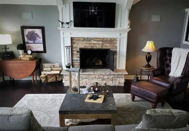 A cozy living room featuring a traditional brick fireplace with a white wood mantel and flat-screen TV mounted above, flanked by a brown leather wingback chair, gray sofas, dark hardwood floors, and warm ambient lighting.