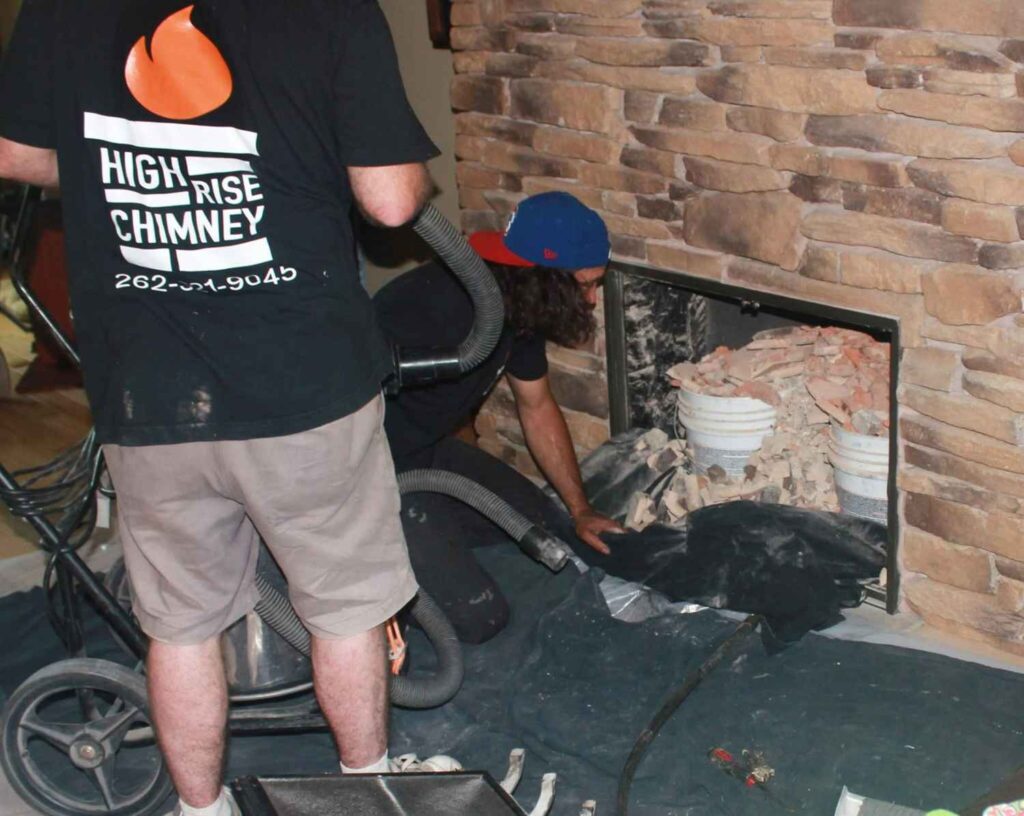 Two High Rise Chimney technicians performing a fireplace cleaning and repair, one using an industrial vacuum hose while the other works inside the firebox removing debris, in front of a stacked stone fireplace surround.