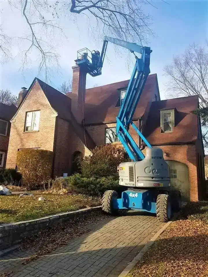 A blue articulating boom lift crane elevated to the roofline of a brick Tudor-style home, with technicians in the bucket performing chimney work on a clear winter day with bare trees.