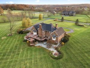 Aerial view of a large, elegant brick mansion surrounded by vast, manicured lawns. The setting is peaceful and expansive, with autumn-colored trees.