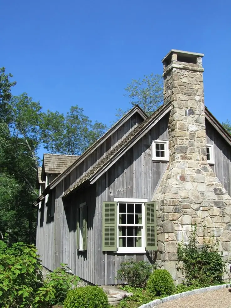 The exterior gable end of a rustic weathered-wood cottage with a large, wide-based fieldstone chimney rising above the roofline, green-shuttered windows, and surrounding trees against a bright blue sky.