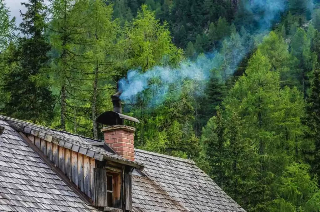 A rustic wooden cabin rooftop with weathered shingle tiles and a red brick chimney emitting a plume of blue-gray smoke, set against a dense forest of tall green conifer trees.