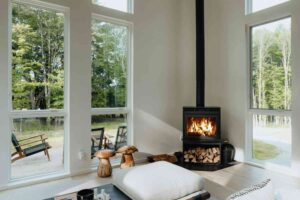 A minimalist, light-filled interior with white walls and large windows overlooking trees, featuring a freestanding black wood-burning stove with a roaring fire, firewood stacked beneath, and simple natural décor including wooden mushroom stools.