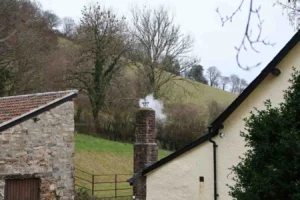 A brick chimney with a chimney pot emitting a thin wisp of white smoke, rising above stone and white-rendered cottage buildings in a rural countryside setting with green hills in the background.
